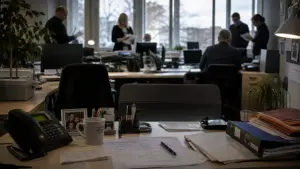 Leerer Arbeitsplatz eines erfahrenen Mitarbeiters im Büro, während Kollegen im Hintergrund Unterlagen prüfen – Sinnbild für stilles Wissen und Wissensverlust im Mittelstand.